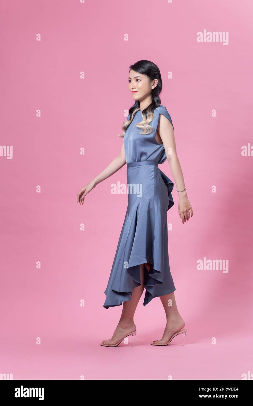 Retrato completo de una mujer joven bonita con vestido azul y tacones altos aislados sobre un fondo rosa Foto de stock