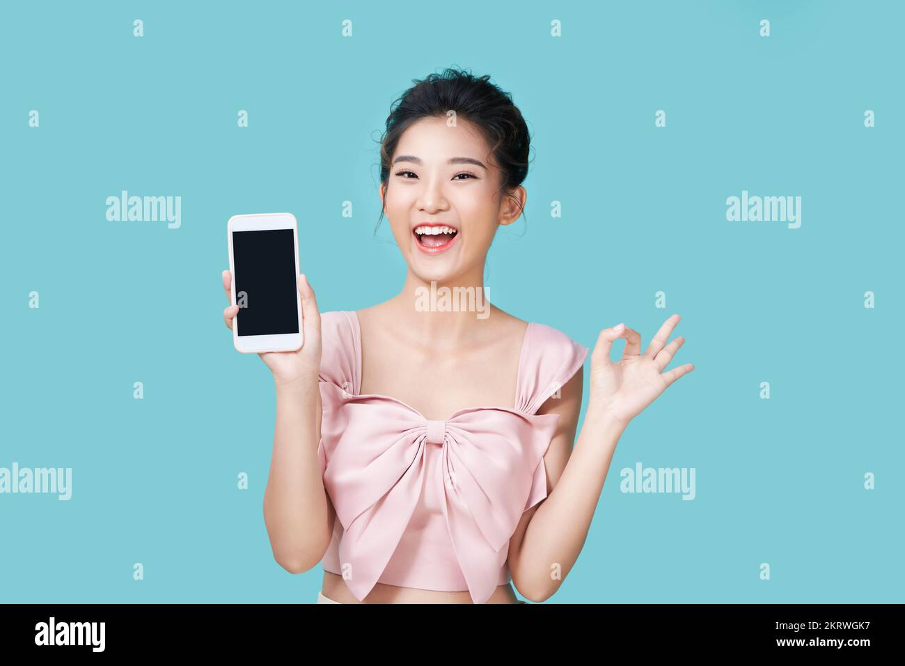 Hermosa mujer asiática sosteniendo smartphone maqueta de pantalla en blanco y muestra el signo ok sobre fondo cian Foto de stock