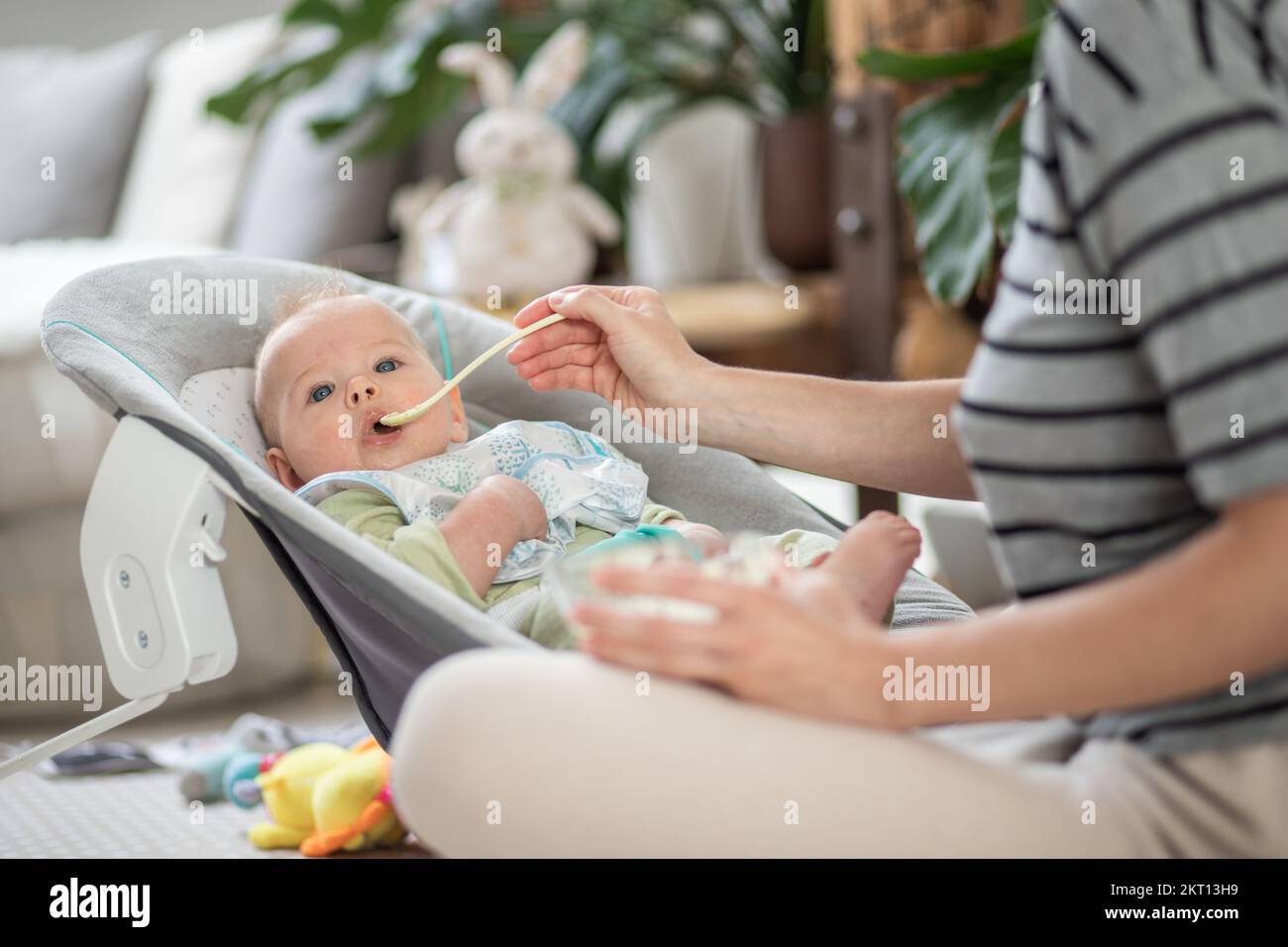 Cuchara madre alimentando a su bebé niño en silla de bebé con puré de frutas. Concepto de introducción de alimentos sólidos para bebés Foto de stock