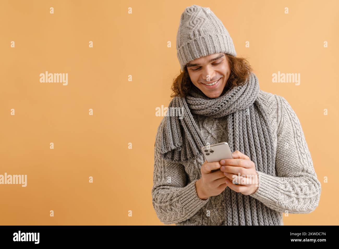 Joven hombre guapo con el pelo largo en invierno sombrero y bufanda sosteniendo el teléfono y mirando en la pantalla sobre fondo marrón aislado Foto de stock