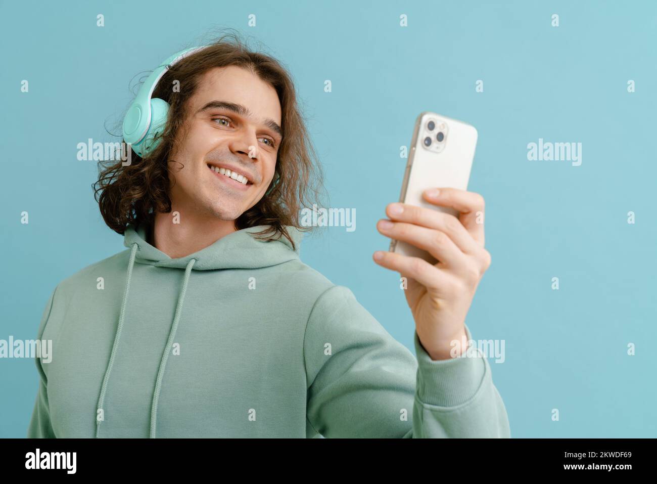 Joven sonriente con el pelo largo en los auriculares sosteniendo el teléfono y mirando la pantalla sobre un fondo azul aislado Foto de stock