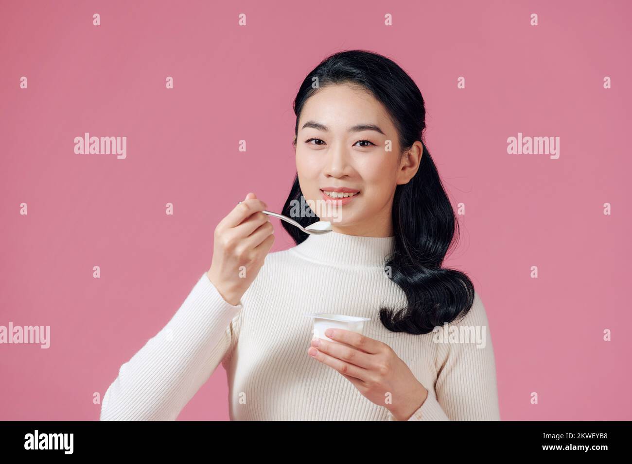 joven hermosa mujer sonriente comiendo yogur fresco sobre rosa Foto de stock