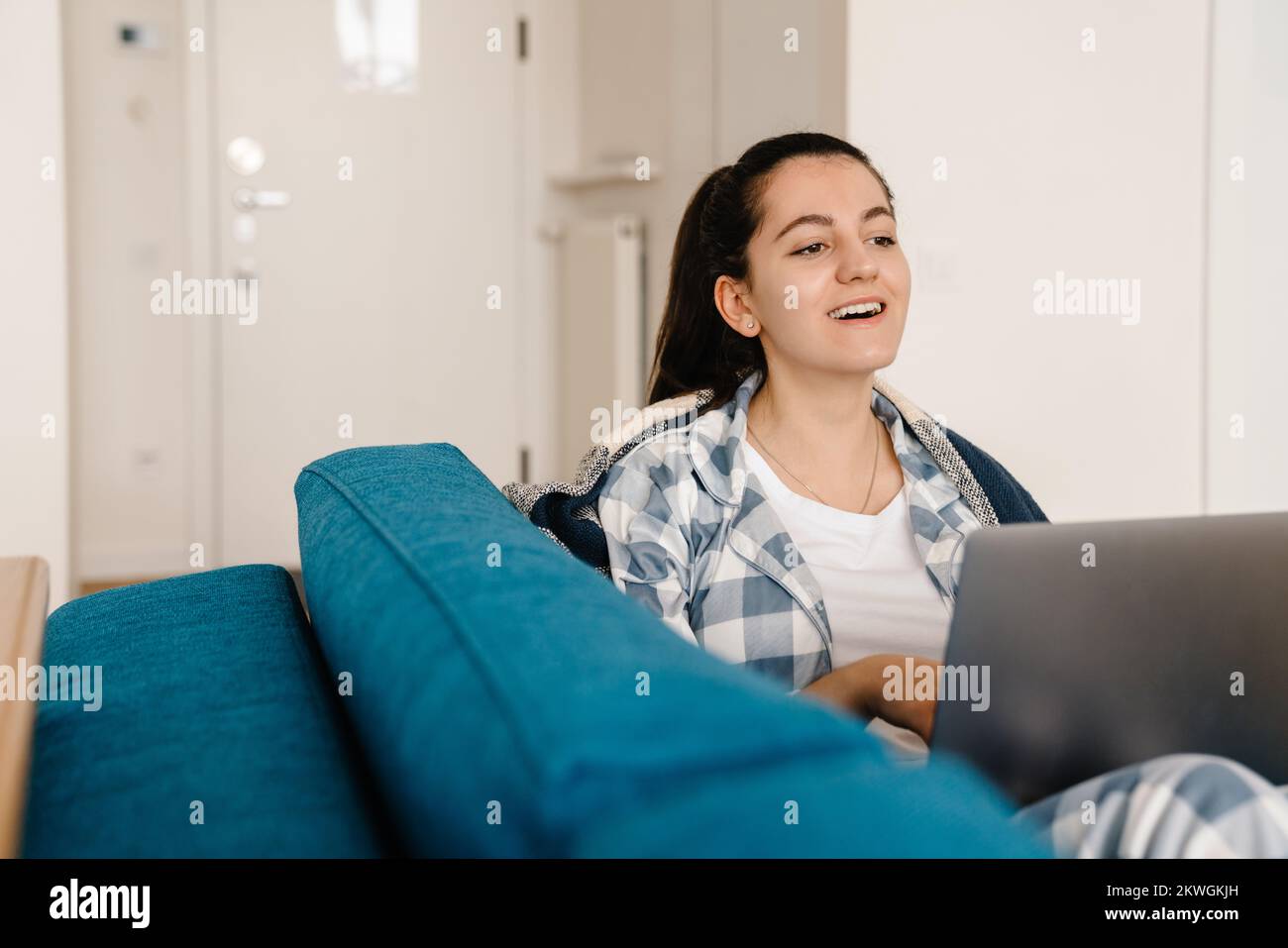 Mujer joven sonriendo y utilizando el ordenador portátil mientras descansa en el sofá de la casa Foto de stock