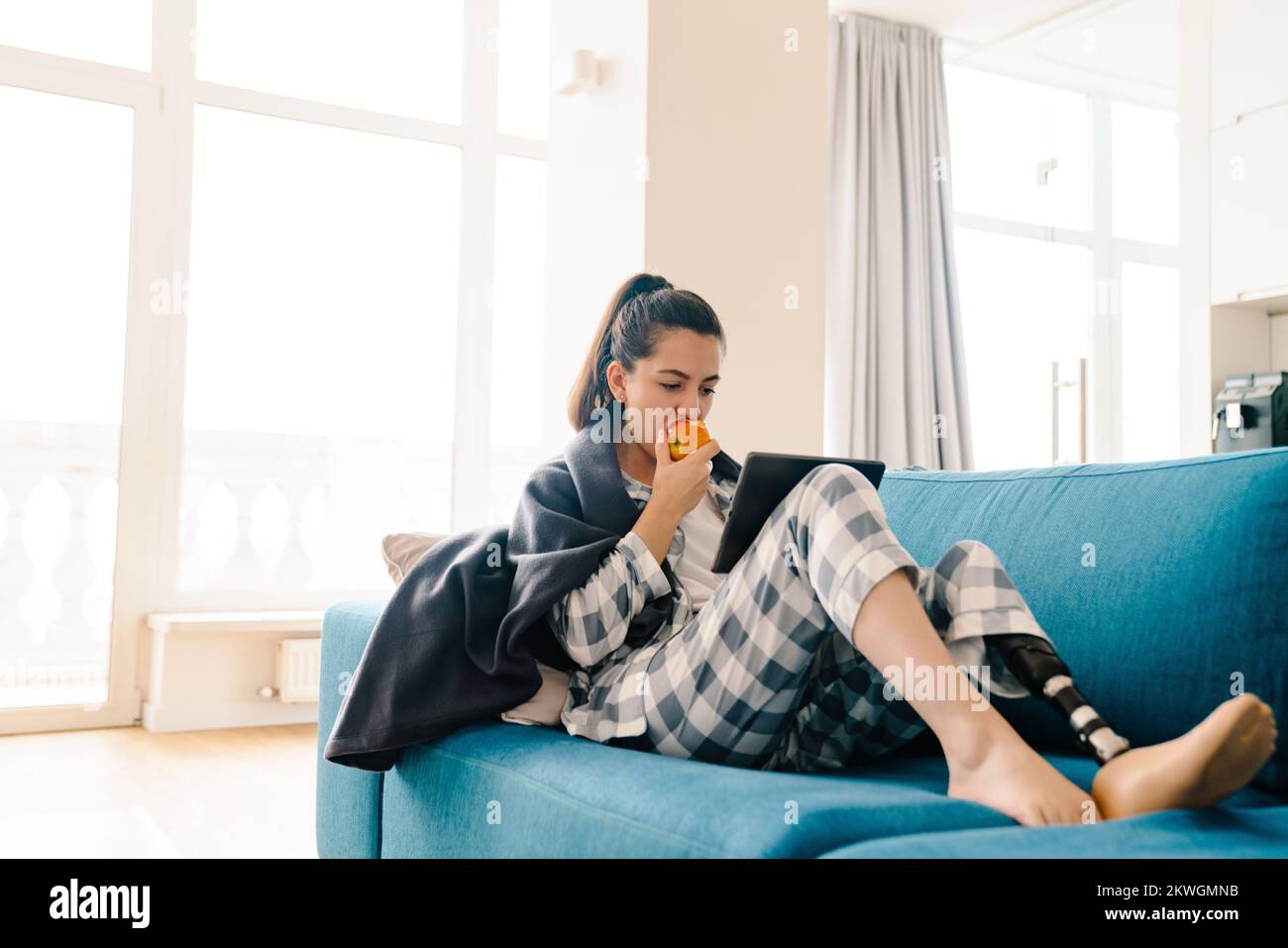 Mujer joven con prótesis usando una computadora tableta y comiendo manzana en el sofá en casa Foto de stock
