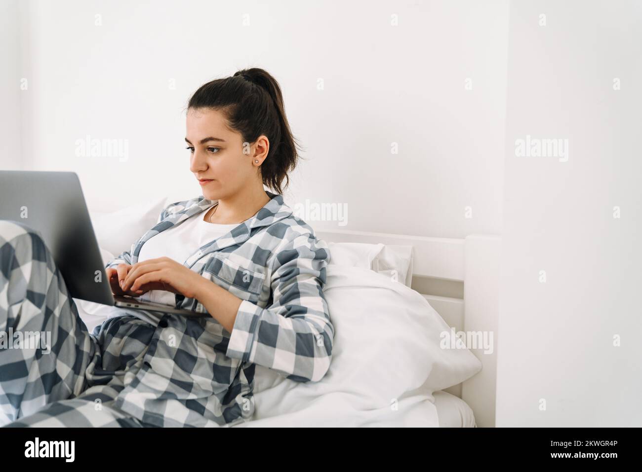 Mujer joven usando pijama usando el ordenador portátil mientras descansa en la cama en casa Foto de stock