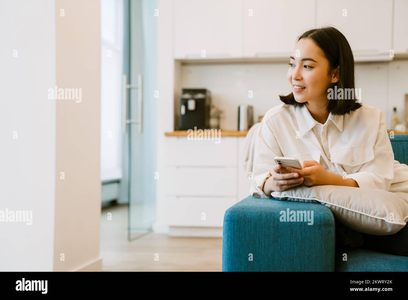 Joven linda chica asiática con teléfono mirando hacia la izquierda sentado en un sofá en un amplio salón acogedor en casa Foto de stock