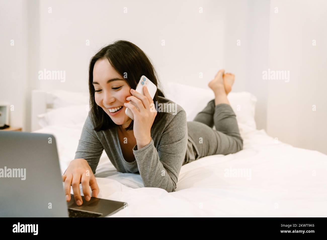 Joven chica asiática sonriente en casa traje liying en la cama con el ordenador portátil y hablando por teléfono en casa Foto de stock