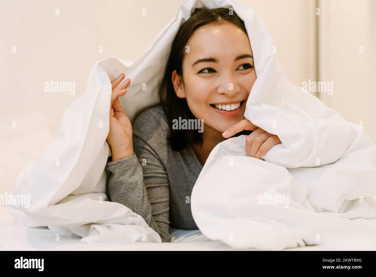 Joven chica asiática sonriente en casa traje tumbado en la cama bajo cubrecamas en casa Foto de stock