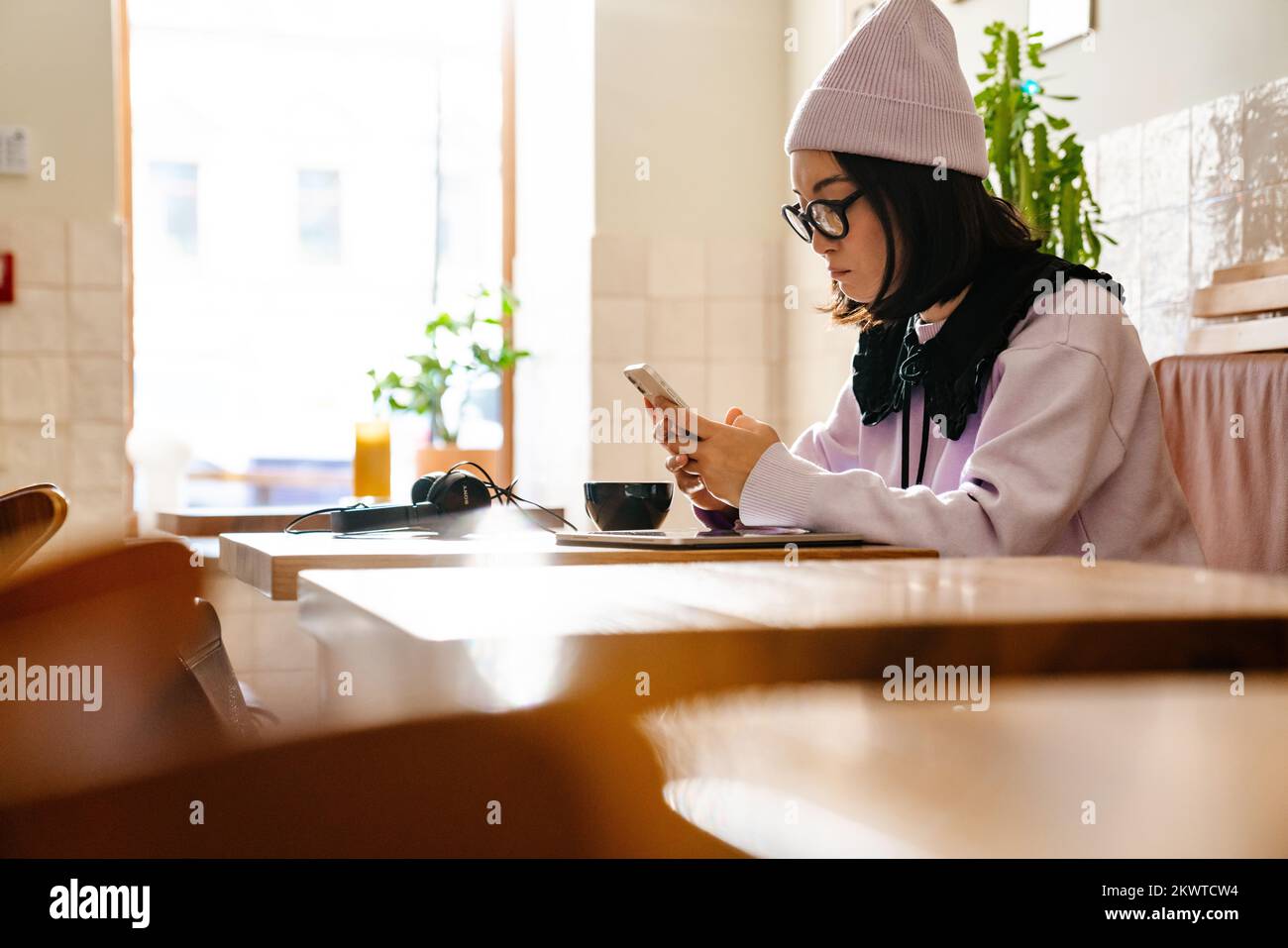 Mujer joven asiática en gafas usando el teléfono celular mientras bebía café en el café Foto de stock