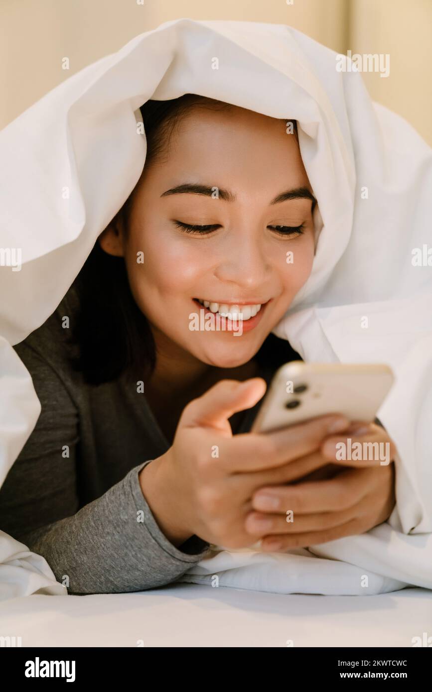 Joven chica asiática sonriente en el hogar traje acostado con el teléfono en la cama debajo de las cubiertas en casa Foto de stock