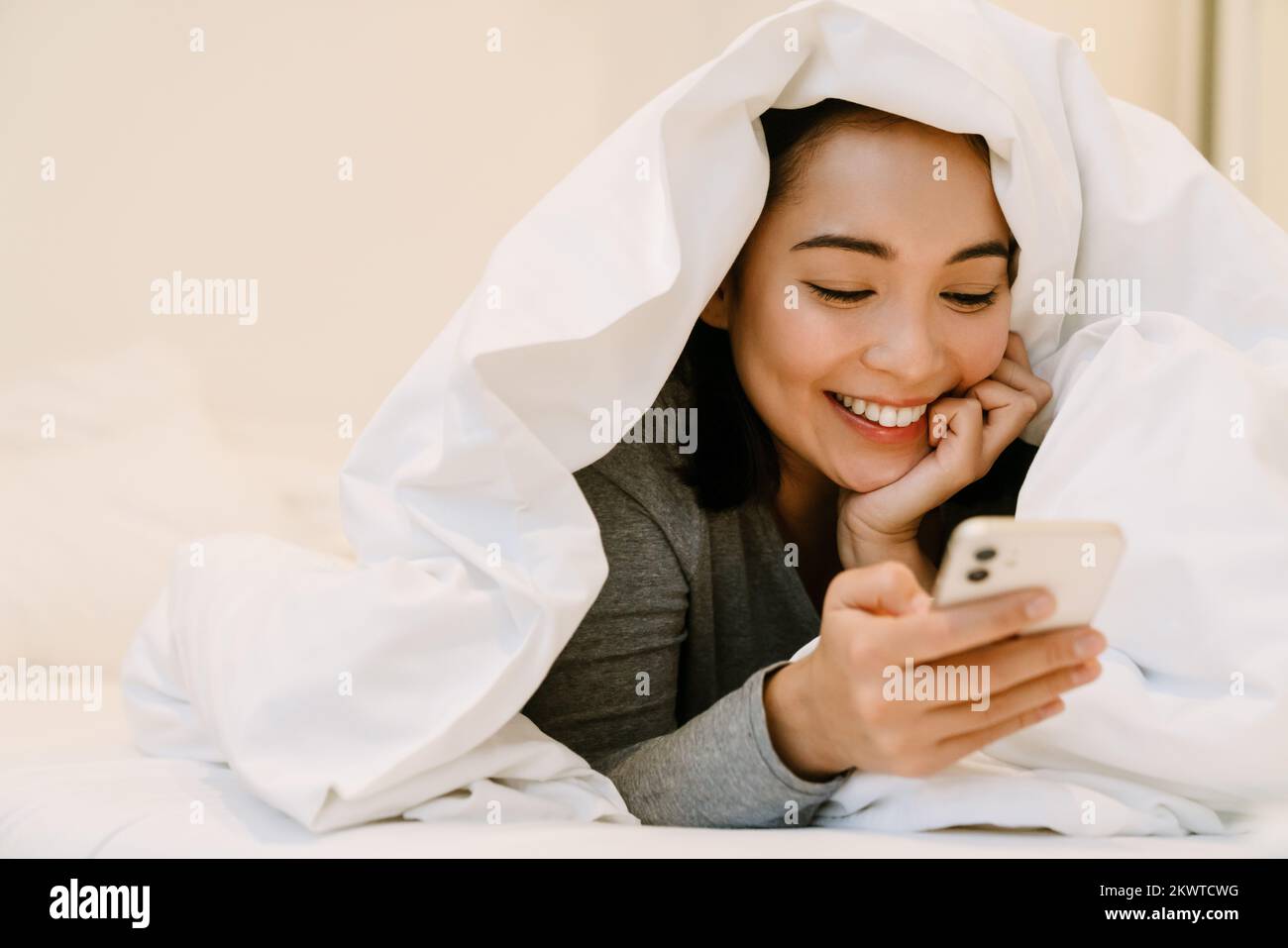 Joven chica asiática sonriente en el hogar traje acostado con el teléfono en la cama debajo de las cubiertas en casa Foto de stock