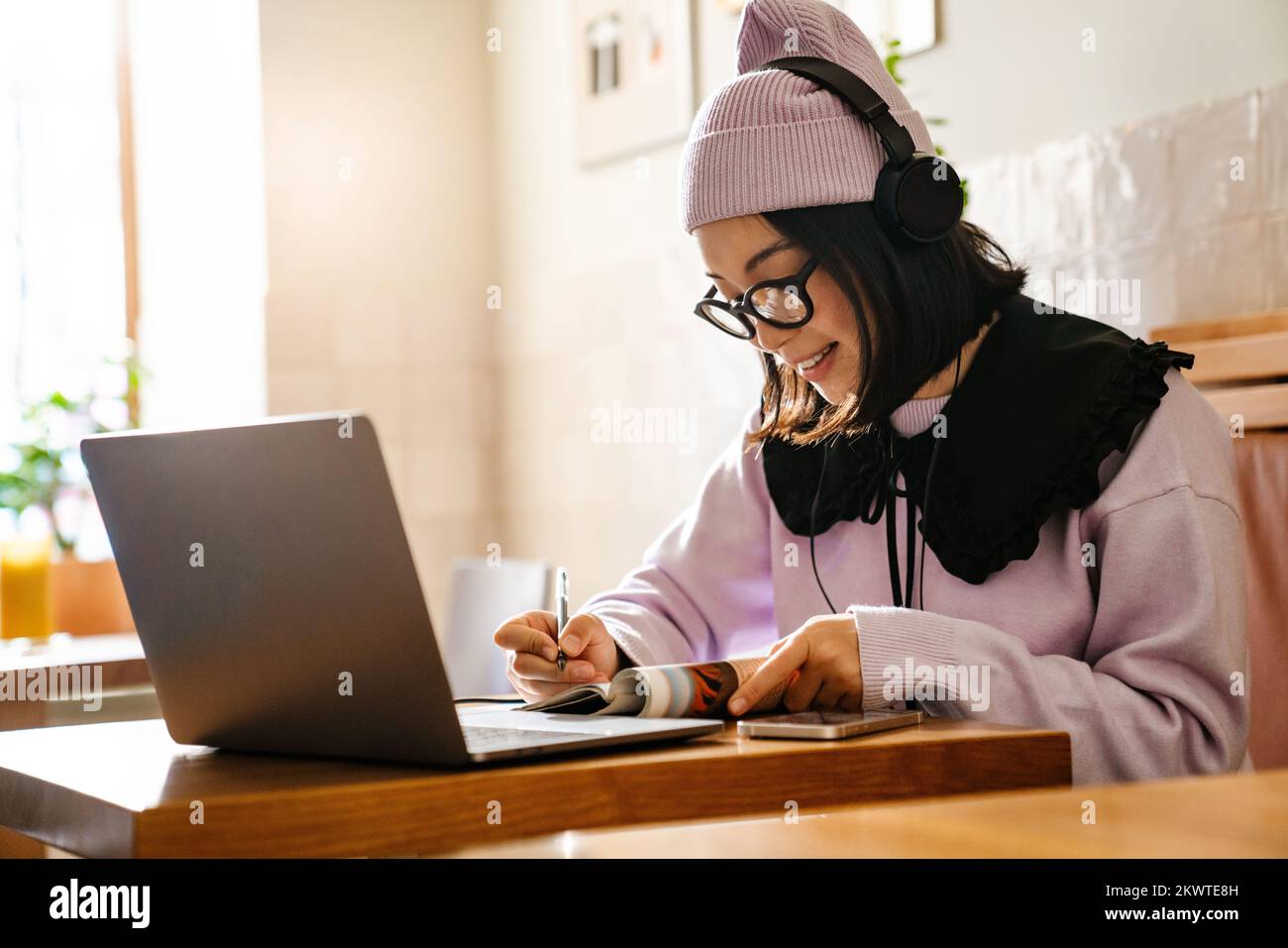 Mujer joven asiática en auriculares haciendo la tarea con el ordenador portátil en el café Foto de stock