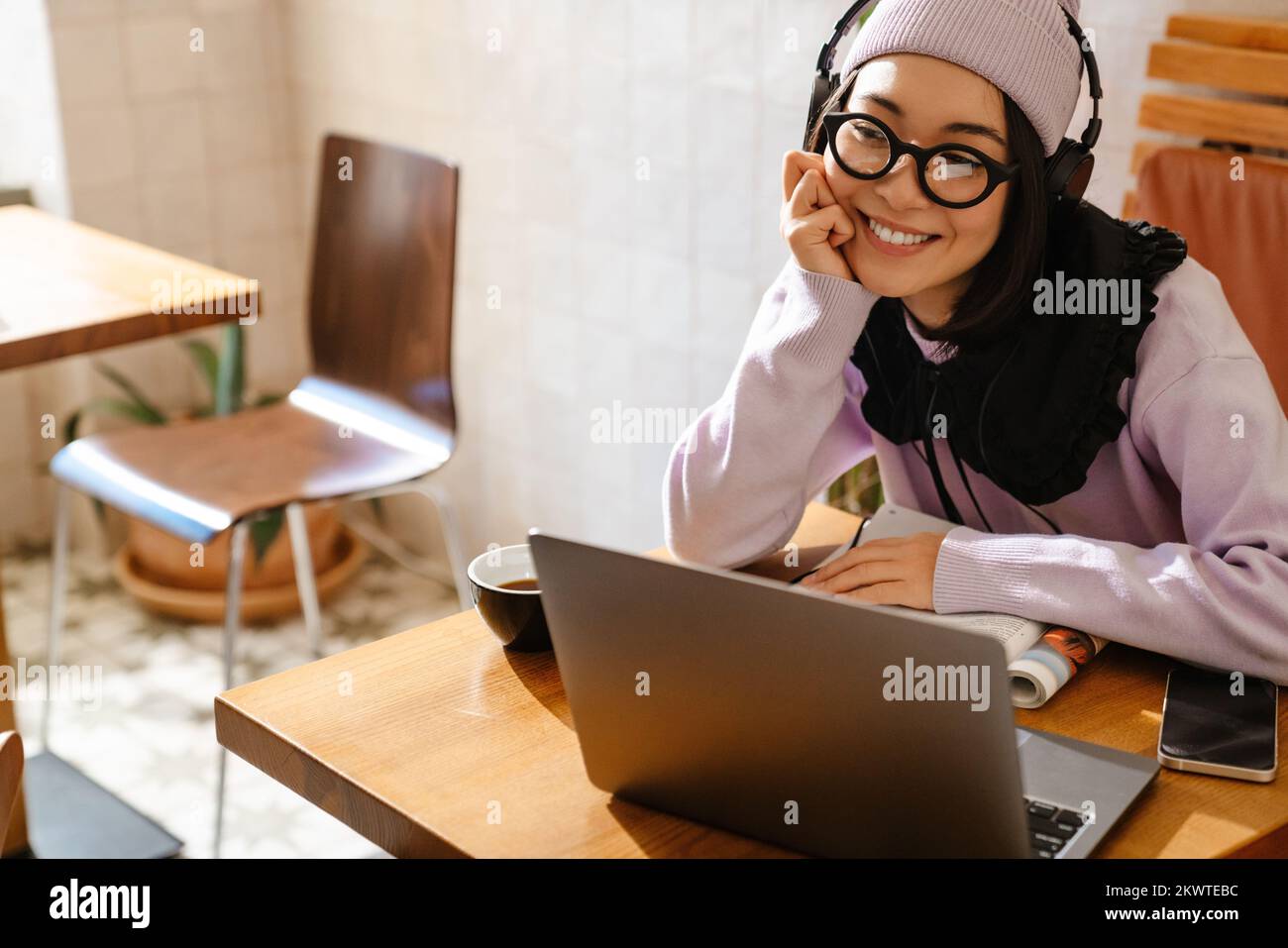 Mujer joven asiática en auriculares haciendo la tarea con el ordenador portátil en el café Foto de stock