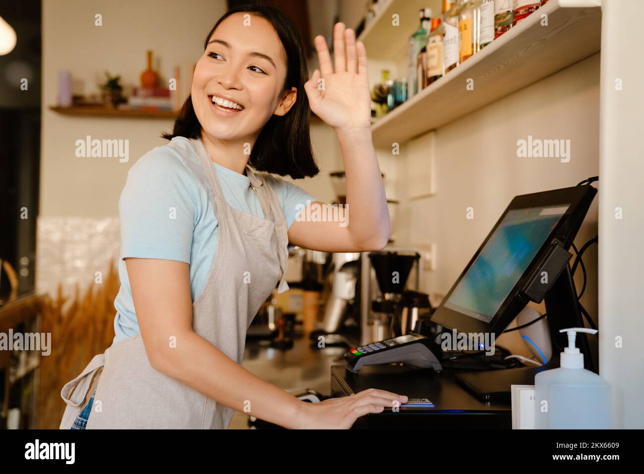 Mujer barista asiática que usa delantal gesturing mientras trabaja con la caja registradora en el café Foto de stock