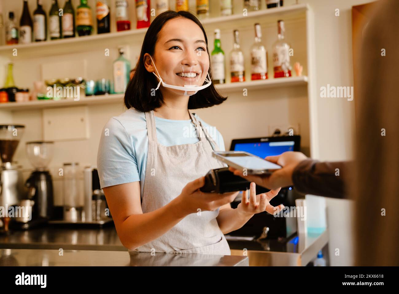 Mujer barista asiática en la cara máscara sonriendo mientras trabaja con el cliente en el café Foto de stock