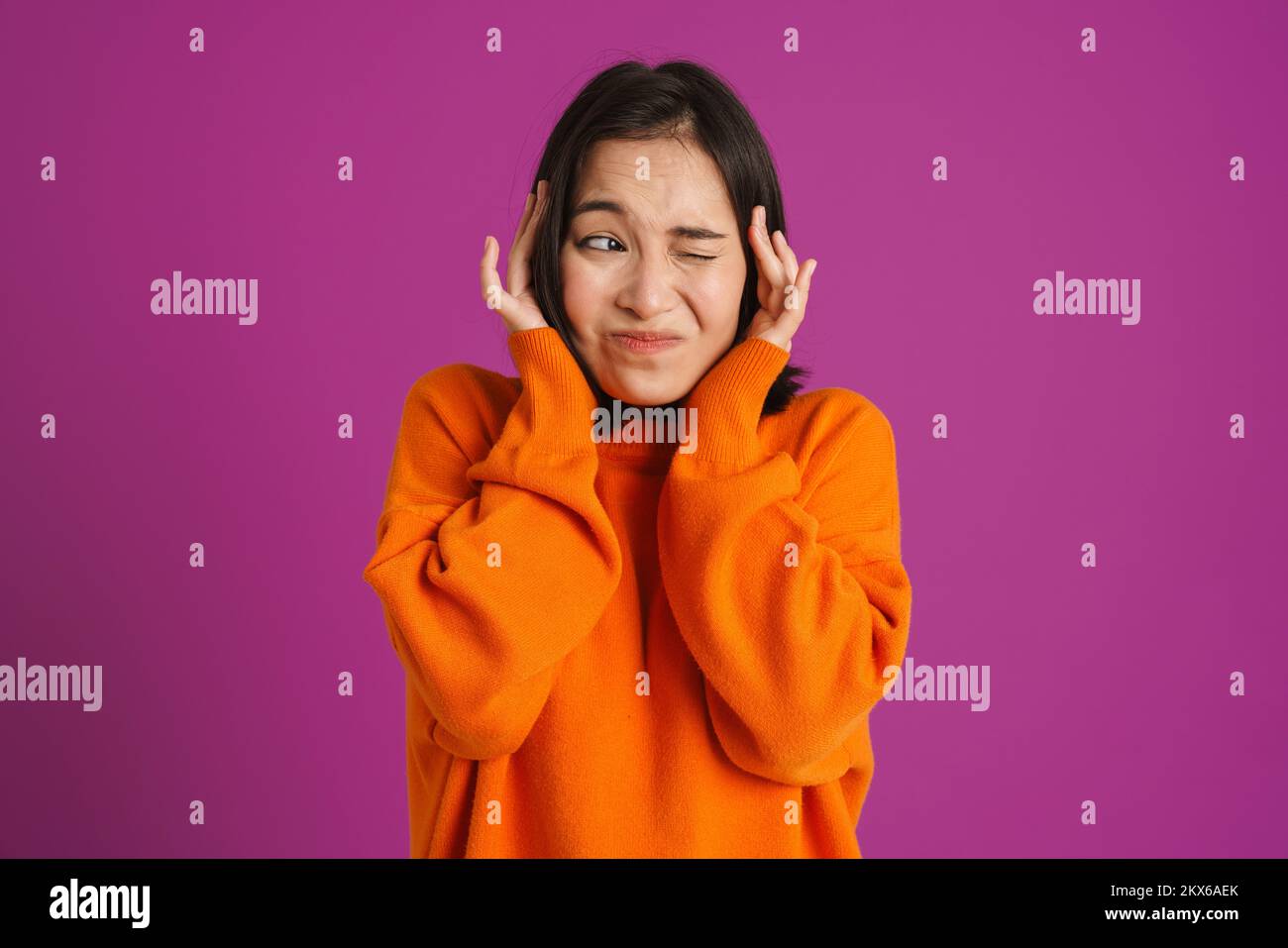 Joven mujer asiática frunciendo el ceño y cubriendo sus oídos aislados sobre fondo púrpura Foto de stock