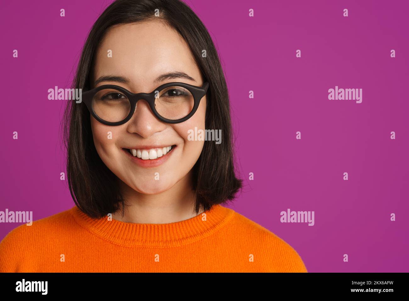 Joven mujer asiática en gafas sonriendo y mirando la cámara aislada sobre fondo púrpura Foto de stock