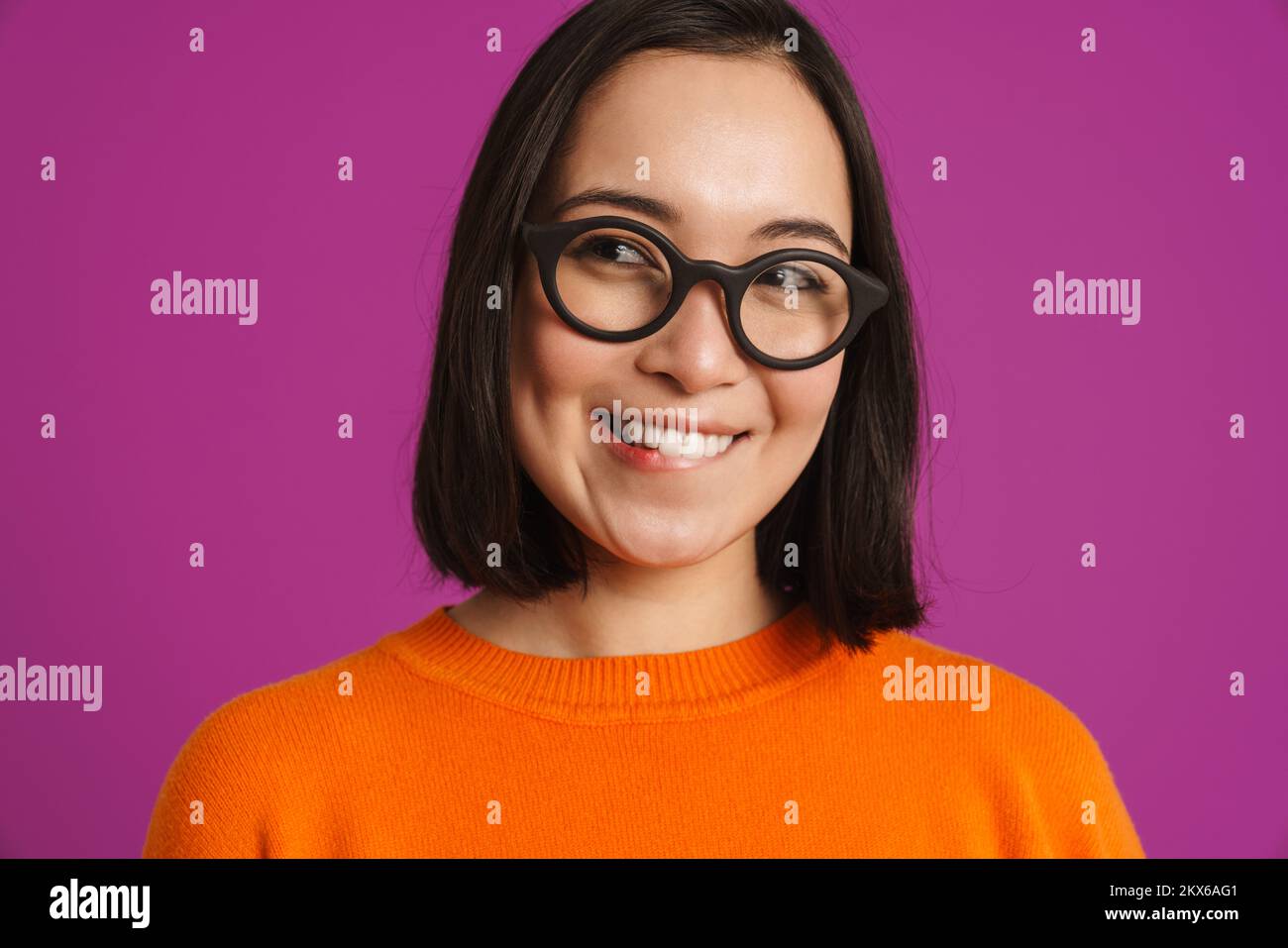Joven mujer asiática en gafas sonriendo y mirando a un lado aislado sobre fondo púrpura Foto de stock