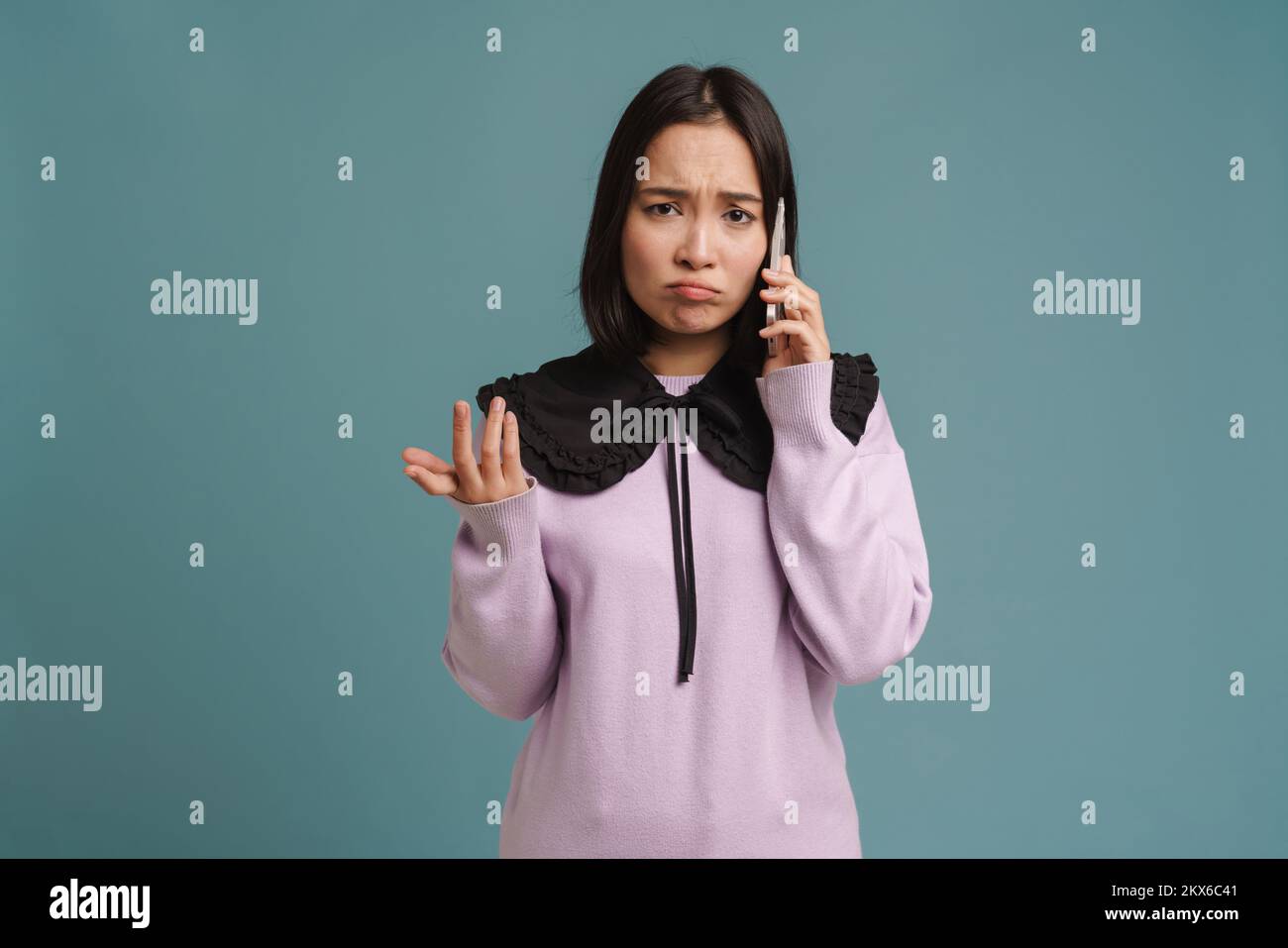 Mujer joven asiática frunciendo el ceño y hablando por teléfono móvil aislada sobre fondo azul Foto de stock