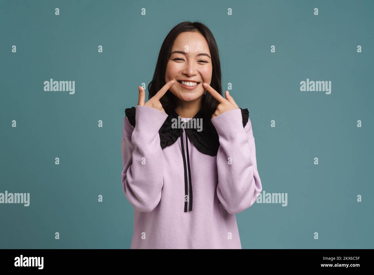 Joven mujer asiática riendo mientras señalaba con los dedos su sonrisa aislada sobre fondo azul Foto de stock