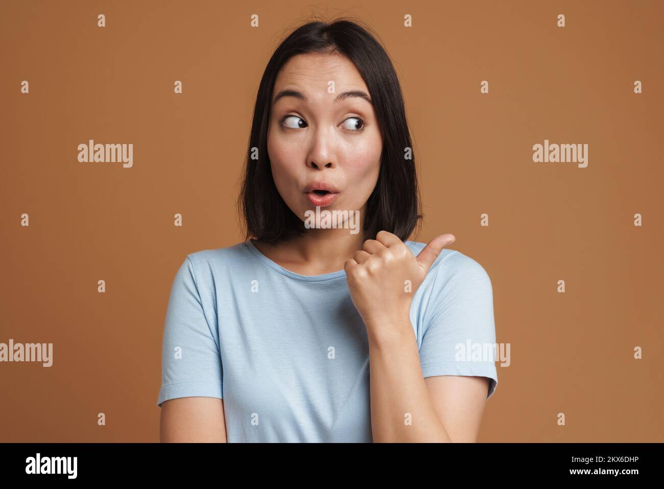 Mujer joven asiática expresando sorpresa y señalando con el dedo a un lado aislado sobre fondo beige Foto de stock