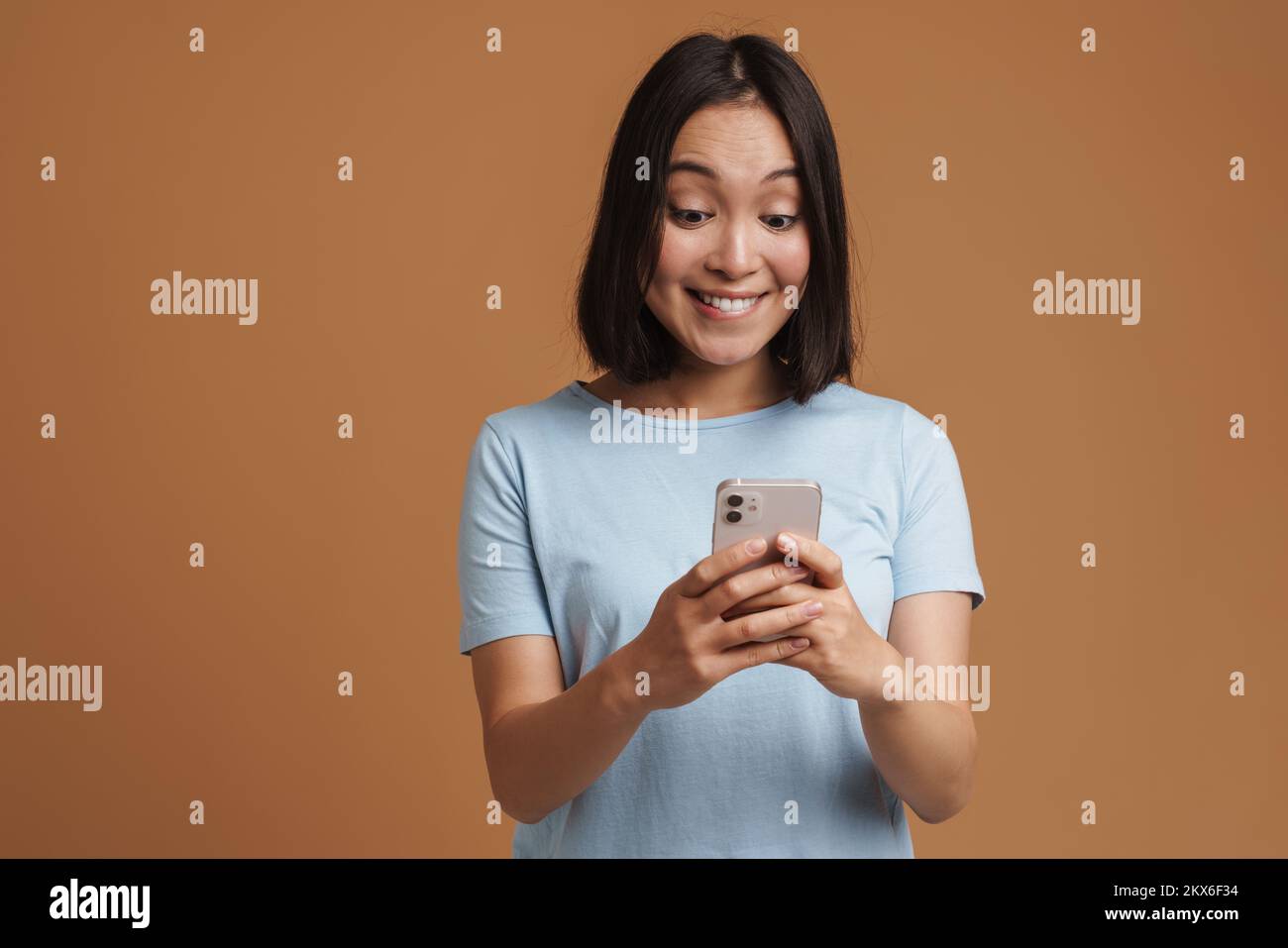Joven asiática con una camiseta que se ríe mientras utiliza un teléfono móvil aislado sobre fondo beige Foto de stock