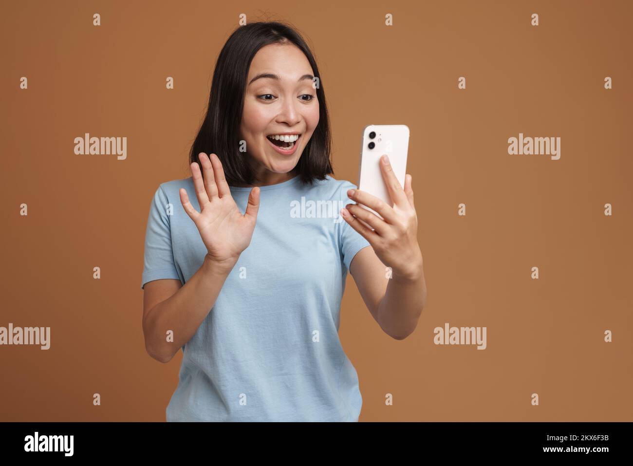 Excitada mujer asiática con camiseta gesturing mientras utiliza el teléfono celular aislado sobre fondo beige Foto de stock
