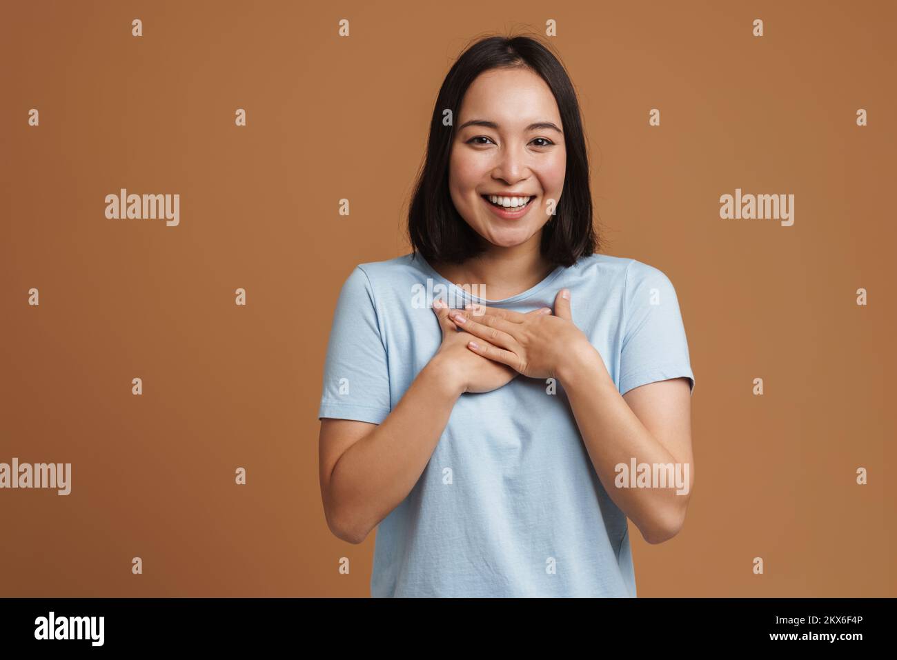 Joven mujer asiática riendo y sosteniendo las manos en su pecho aislado sobre fondo beige Foto de stock