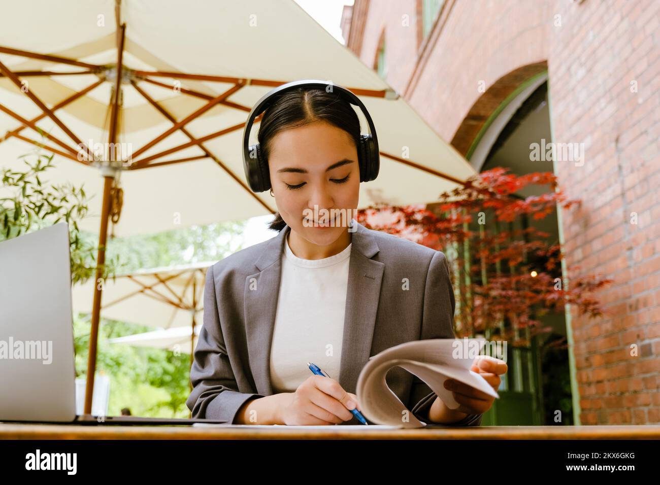 Mujer asiática en auriculares escribiendo notas mientras trabaja con el portátil en el café al aire libre Foto de stock