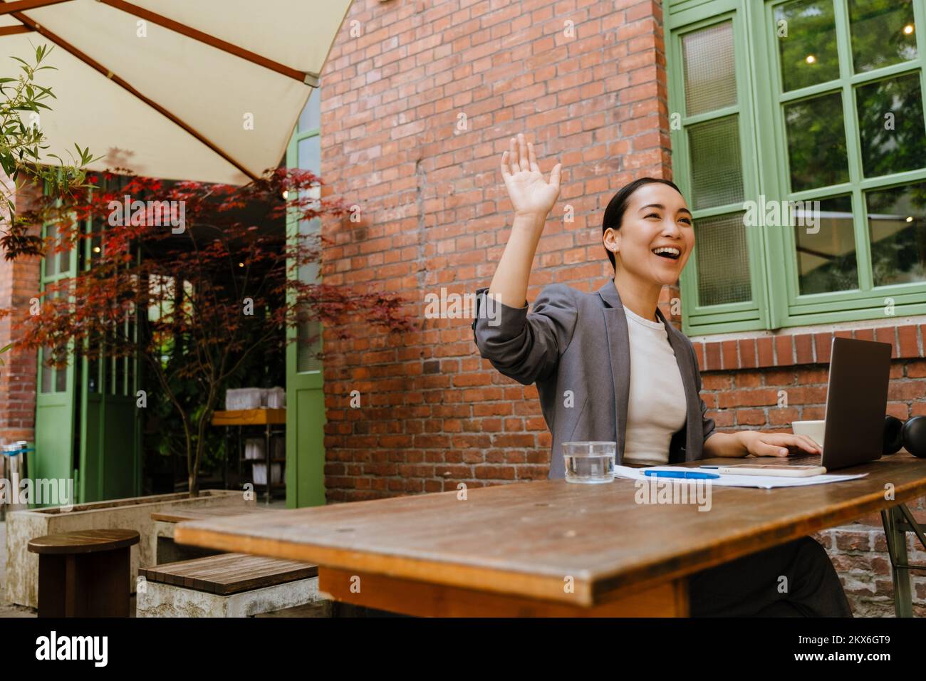 Mujer joven asiática haciendo gestos y trabajando con el ordenador portátil mientras se sienta en el café al aire libre Foto de stock