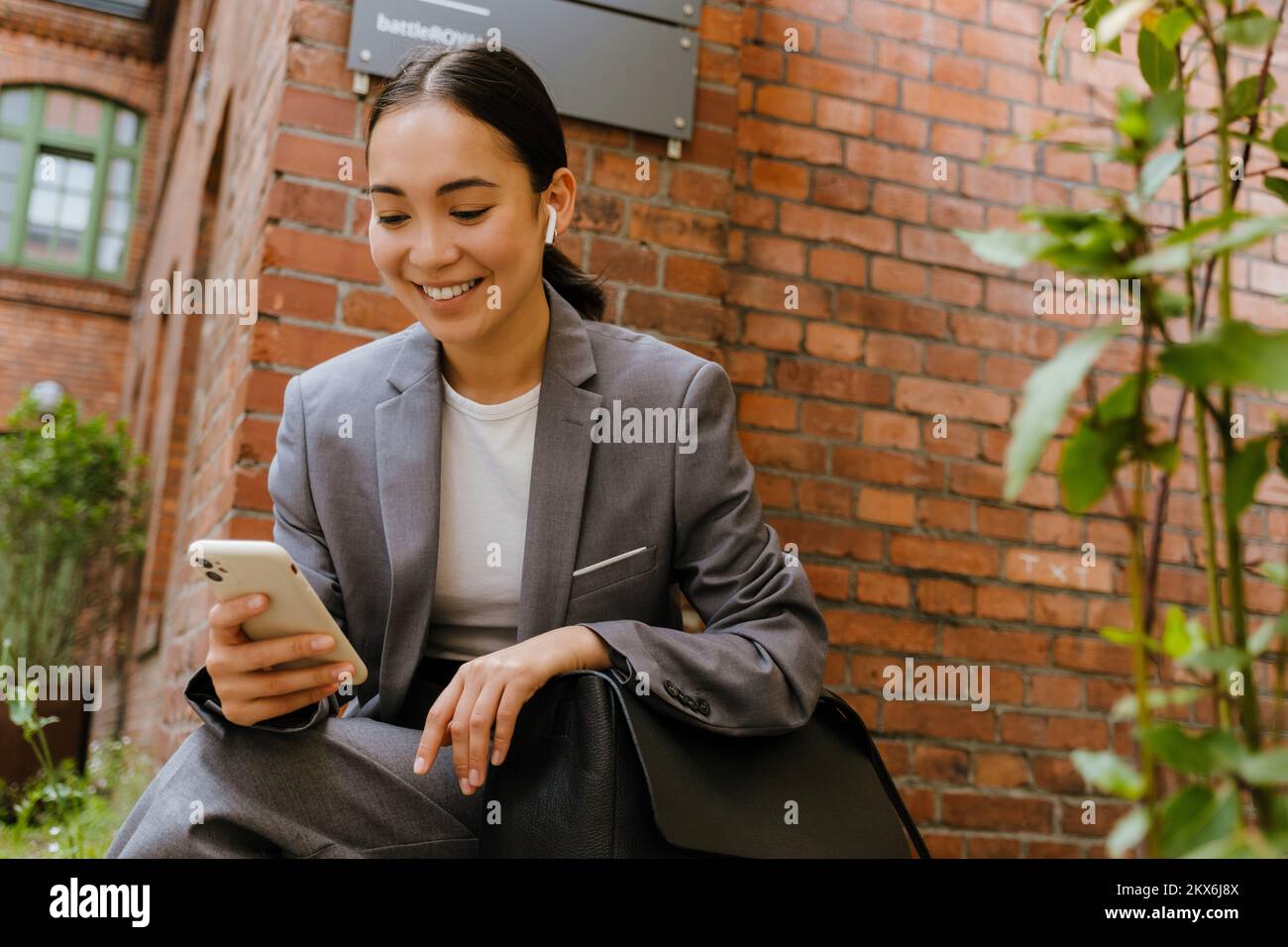 Mujer asiática en auriculares usando el teléfono móvil mientras se sienta en el edificio al aire libre Foto de stock
