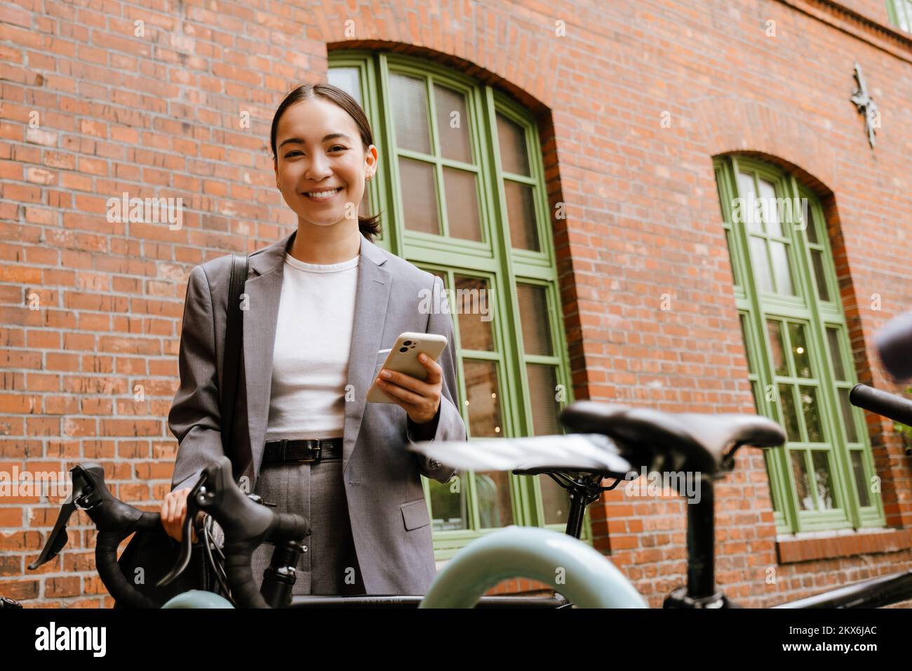 Joven empresaria asiática sonriendo y utilizando el teléfono móvil mientras está de pie en bicicleta en la calle Foto de stock