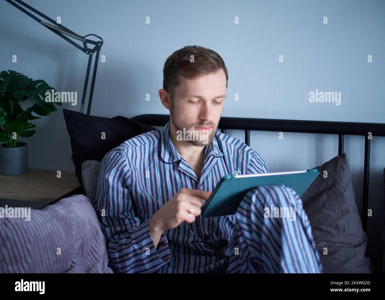 Atractivo caucásico barbudo con ropa de dormir usando una tableta de ordenador sentado en por la mañana educando, trabajando o haciendo compras en línea en Internet. Hombre usando una