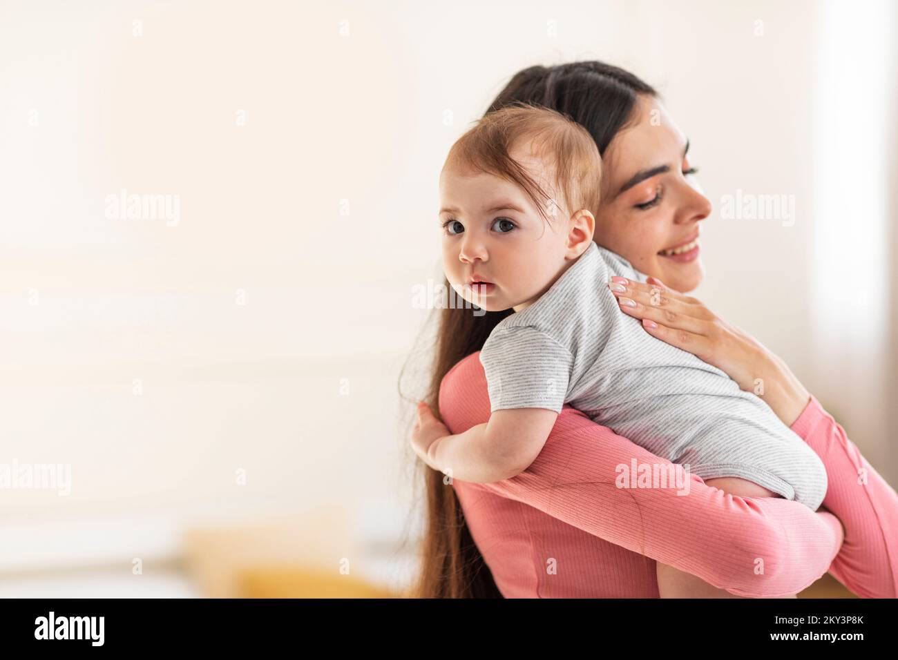 Joven mamá sosteniendo y abrazando al adorable bebé, disfrutando de la maternidad y el cuidado de los niños, de pie en la habitación en casa Foto de stock