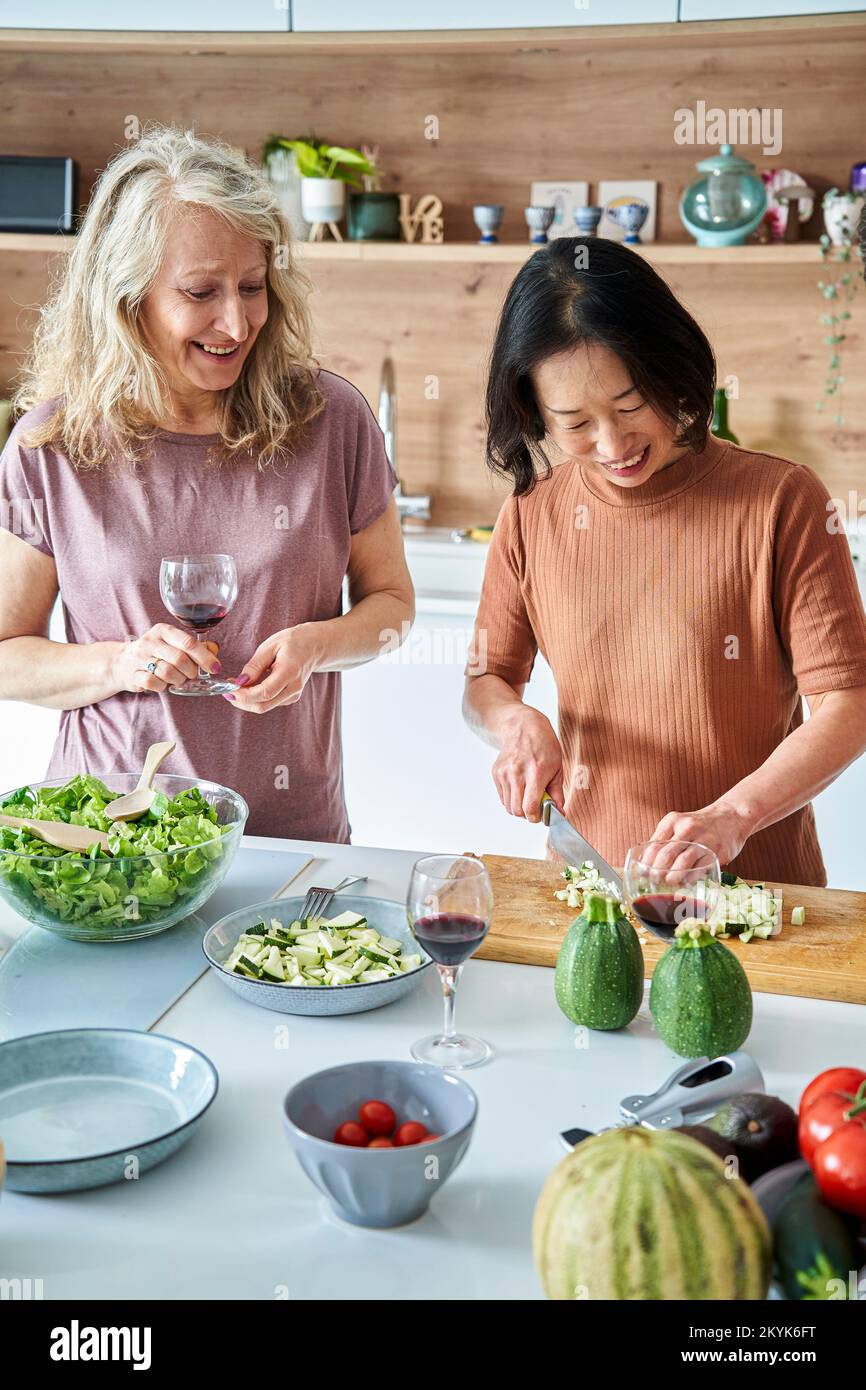 Mujer asiática senior cortando verduras mientras bebe vino con un amigo Foto de stock