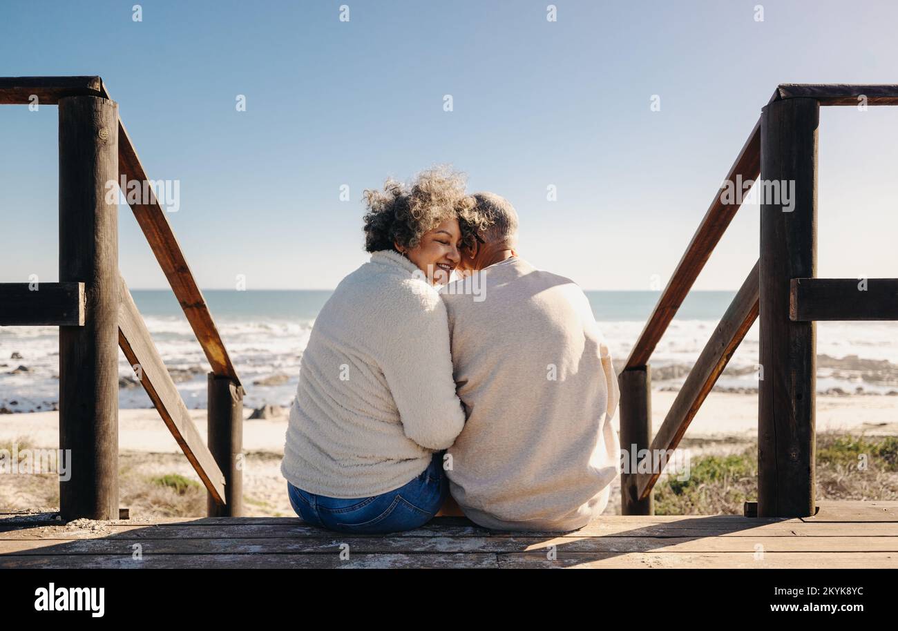Mujer joven romántica sonriendo alegremente mientras se sienta en un puente de madera junto al mar con su marido. Jubilados pareja de ancianos pasar algún tiempo de calidad tog Foto de stock
