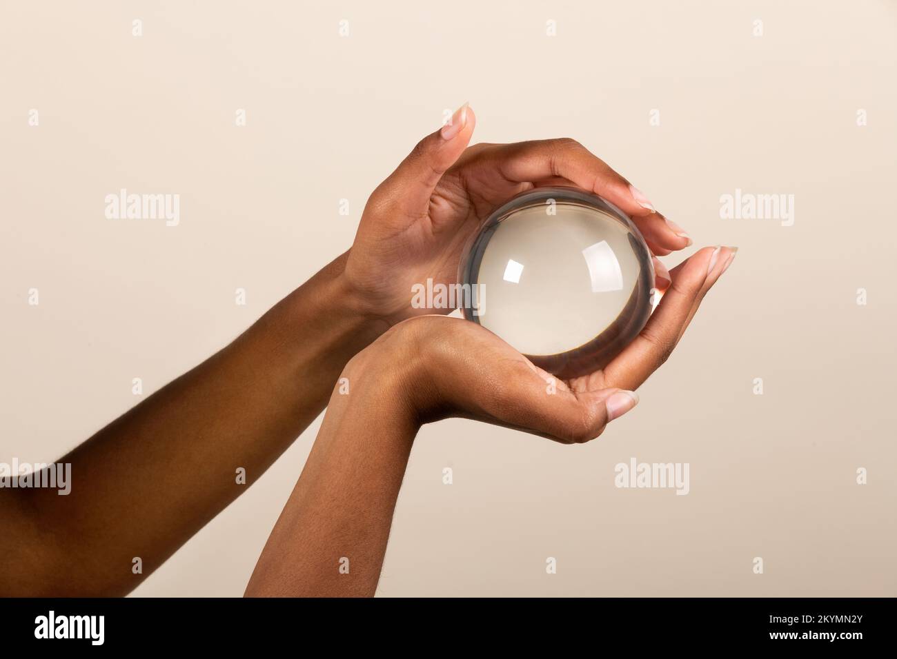 Manos de mujer negra sosteniendo una bola de cristal transparente con un ligero reflejo sobre fondo beige Foto de stock