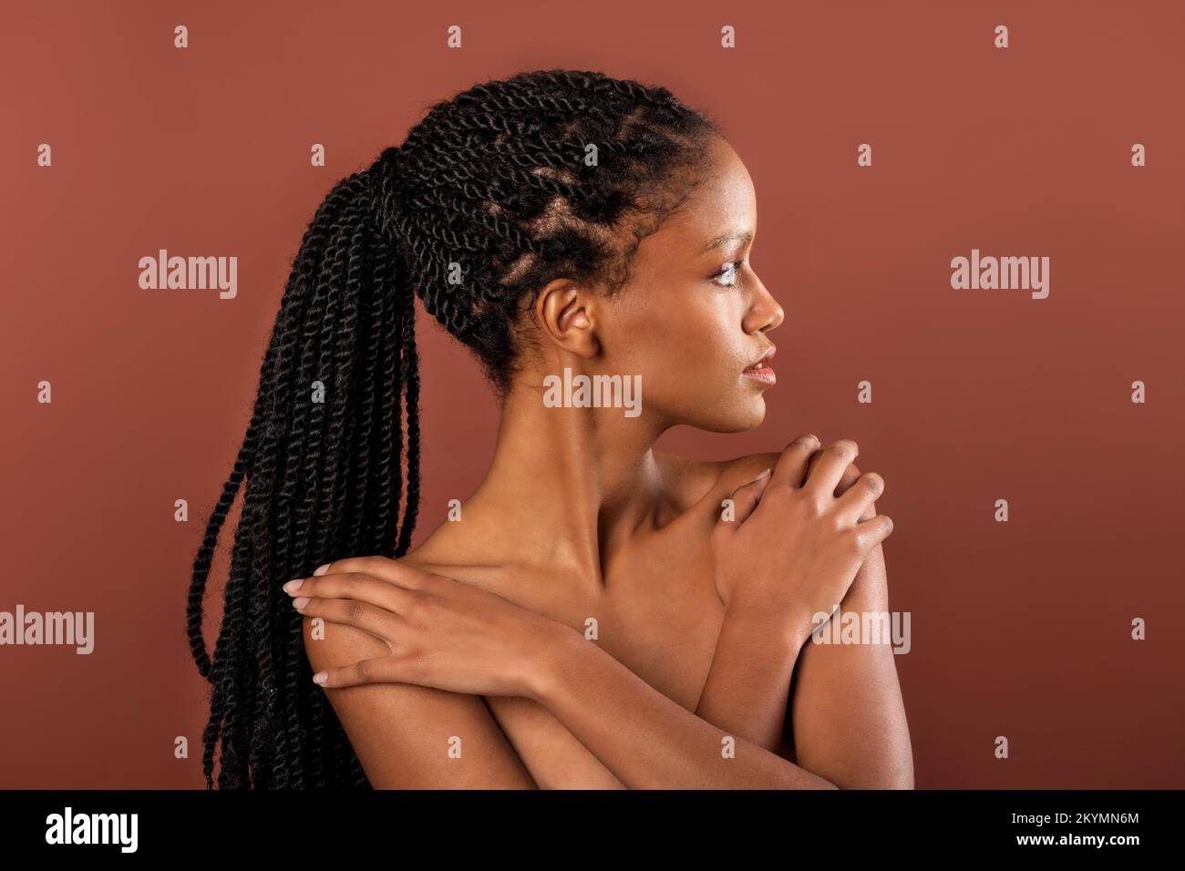 Preciosa joven mujer étnica con trenzas afro sosteniendo las manos sobre hombros descalzos mirando sobre fondo marrón Foto de stock