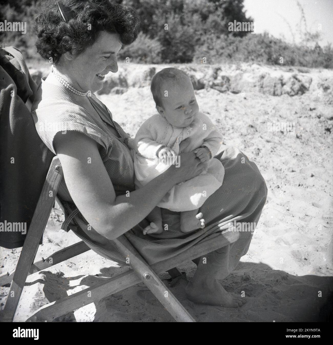 1950s, histórico, fuera en una playa arenosa, una feliz madre sentada en una tumbona sosteniendo a su niño en su regazo, Inglaterra, Reino Unido. Foto de stock