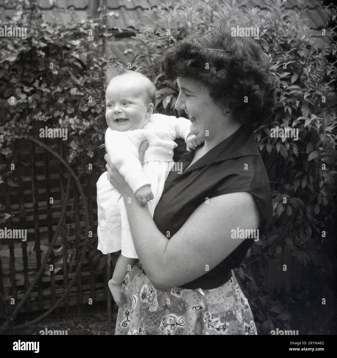 1950s, histórico, fuera en un jardín, una madre feliz sosteniendo a su bebé sonriente, Inglaterra, Reino Unido. Foto de stock