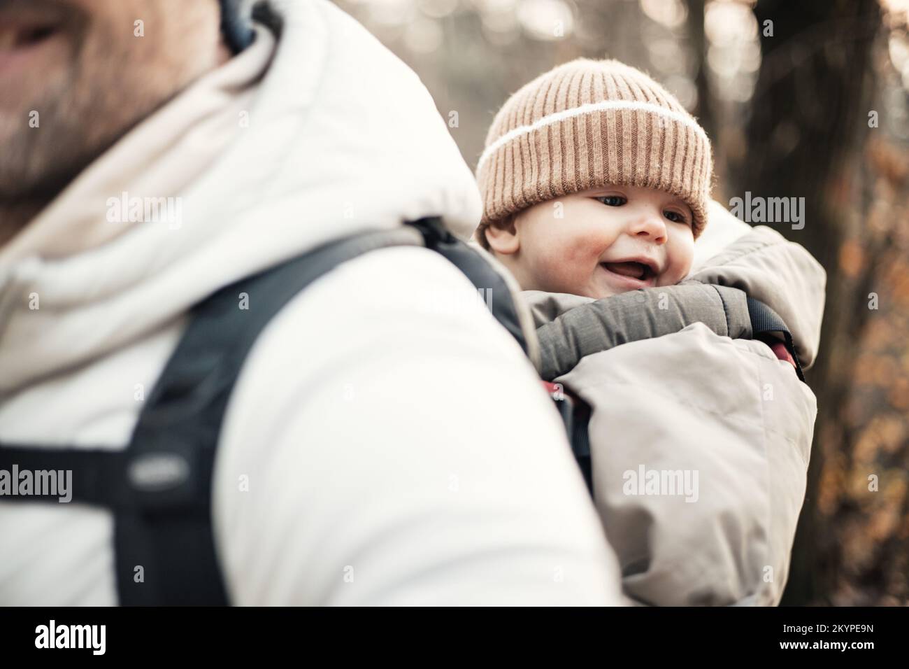 Padre deportivo llevando a su hijo pequeño con traje de mono de invierno y gorra en mochila de senderismo en el bosque de otoño Foto de stock