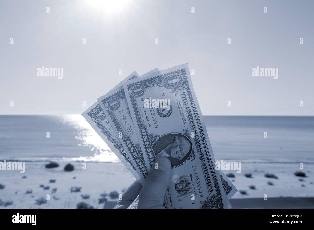 Persona que tiene tres billetes de un dólar en la mano en el fondo del mar, la playa de arena y el cielo en el soleado día de verano. Concepto de dinero, finanzas, dinero, viajes, turismo, vacaciones, descanso. Color azul Foto de stock