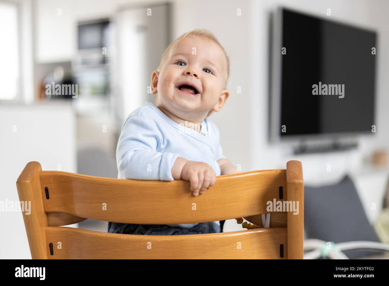 Bebé feliz sentado en una silla alta de madera de diseño escandinavo tradicional y riendo a todo volumen en un hogar moderno y luminoso. Sonrisa linda del bebé Foto de stock