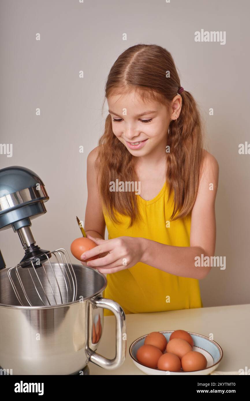 Una chica positiva de 9-10 años cocina pastel casero en la cocina, bate los huevos en una batidora en la mesa de la cocina. Contra el fondo de un blanco-gris Foto de stock