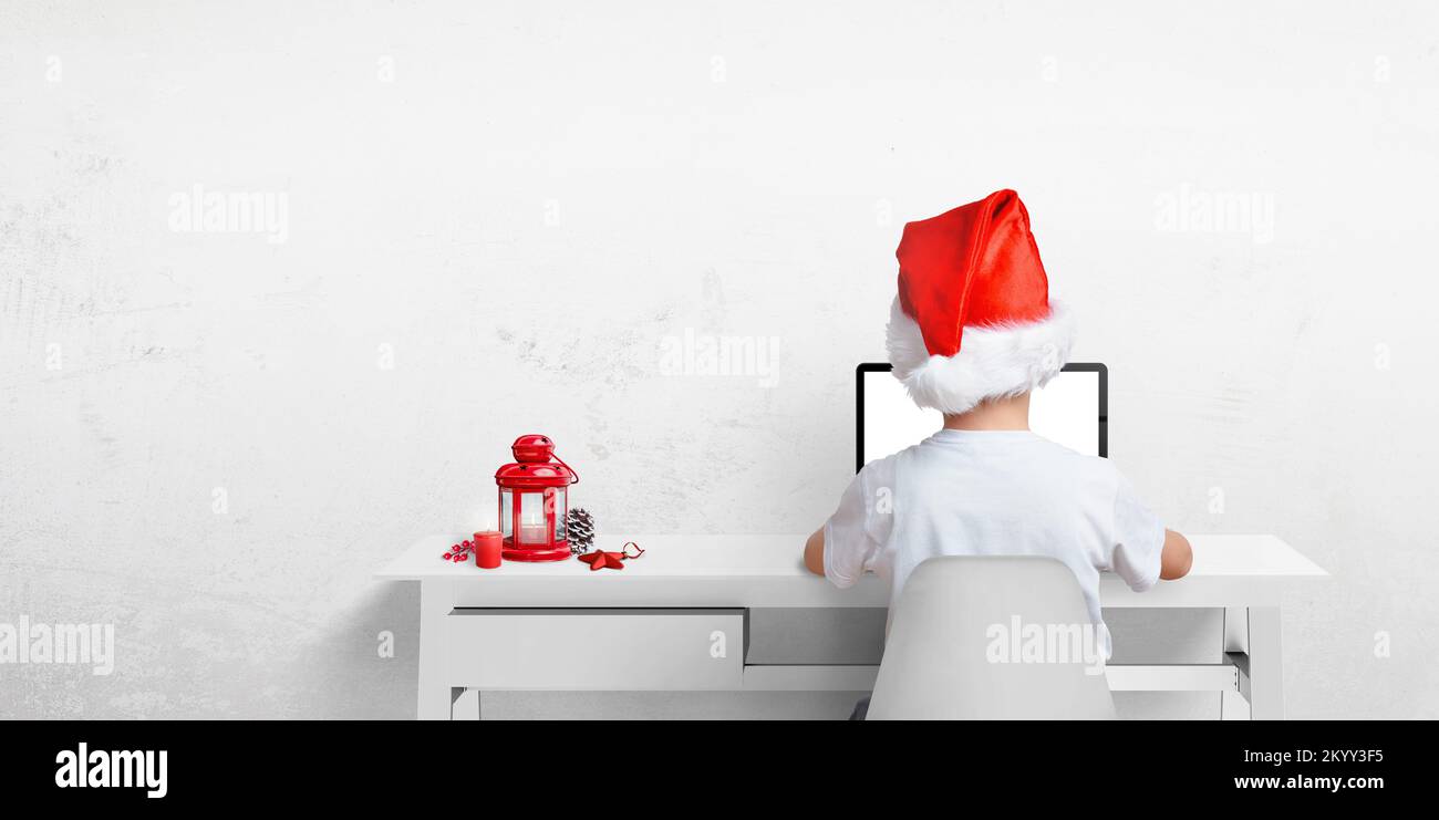 Niño trabajo en ordenador portátil con sombrero de Navidad de Papá Noel. Linterna roja con decoración en el escritorio. Espacio de copia Foto de stock