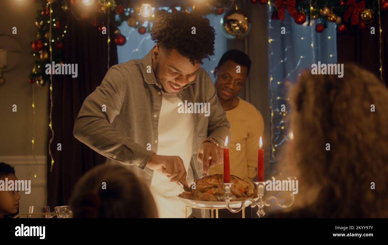 Hombre afroamericano corta pavo o pollo con cuchillo. Amigos multiculturales que celebran Navidad o Año Nuevo. Servido mesa de vacaciones con velas. Ambiente cálido de la cena de Navidad en casa. Foto de stock