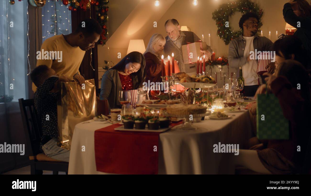 Feliz familia multicultural celebrando Navidad o Año Nuevo 2023, intercambiando regalos. Servido mesa de vacaciones con diferentes platos y velas. Ambiente cálido de la cena de Navidad familiar en casa. Foto de stock