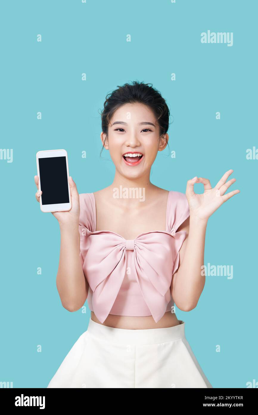 Alegre mujer asiática sosteniendo un smartphone y mostrando el signo ok sobre un fondo cian claro. Foto de stock