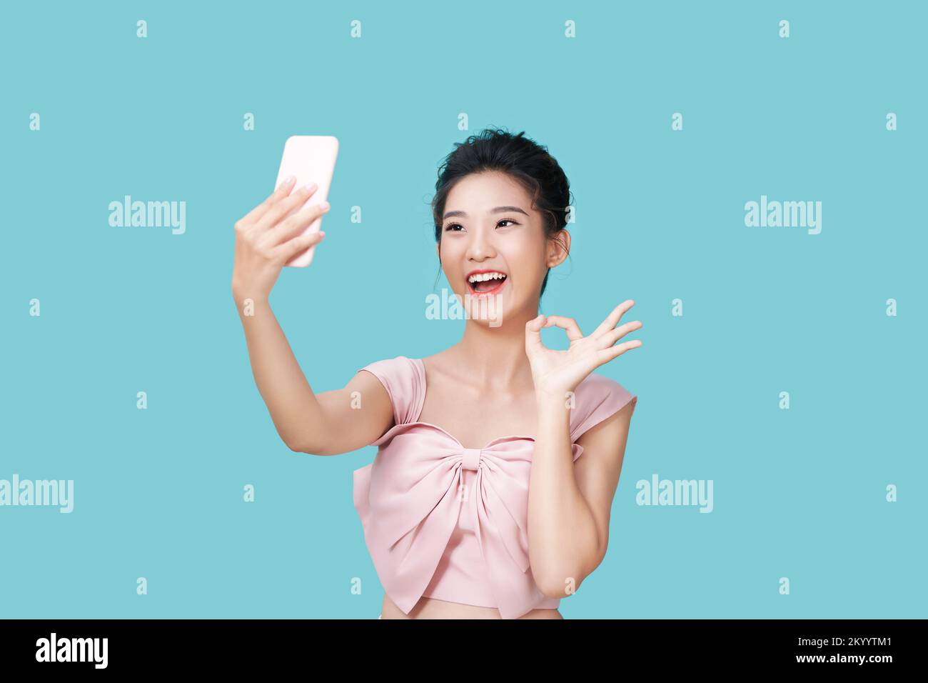 Mujer joven feliz haciendo caras tontas mientras toma selfie con la aplicación para smartphone con filtros, de pie sobre fondo azul Foto de stock