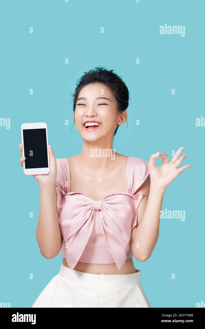Alegre mujer asiática sosteniendo un smartphone y mostrando el signo ok sobre un fondo cian claro. Foto de stock